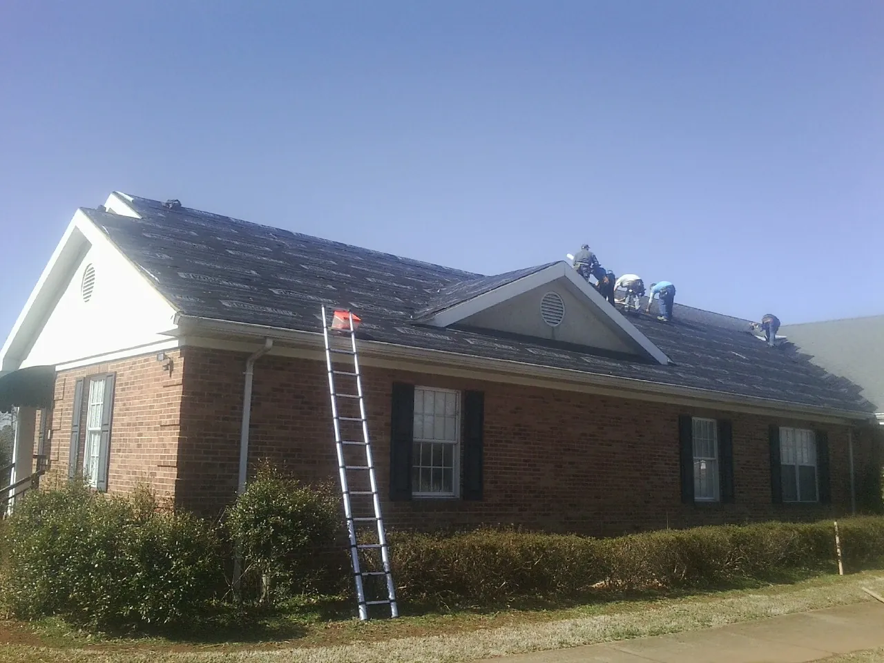 Crew tearing shingles off a brick ranch roof with a ladder set — Metal Roof Repair work by our crew in Minco
