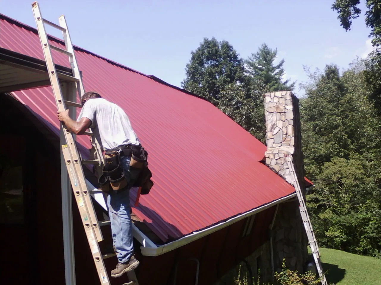 Roofer on a ladder beside a red metal roof and stone chimney — Soffit & Fascia Repair by Roofing Brothers in Minco